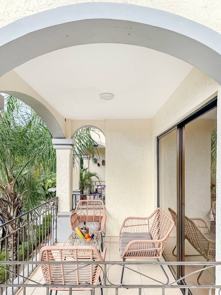 Covered balcony with wicker chairs and a small table, sliding glass door and palm trees beyond the railing
